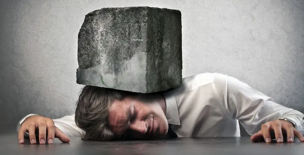 A man is asleep on a table with a large rock on his head.